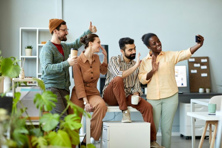 Side view of four young cheerful employees waving hands to smartphone camera